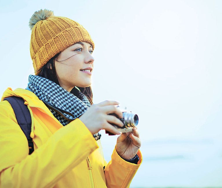 Woman wearing a hat coat and scarf is taking pictures of her surrounding landscape.