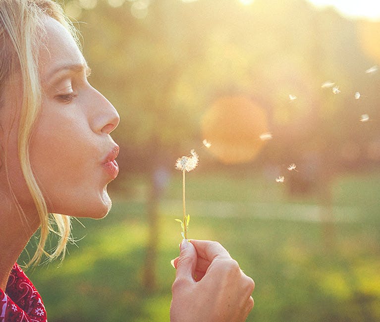 Eine Frau bläst auf eine Pusteblume, denn ihr Mittel gegen Heuschnupfen wirkt.