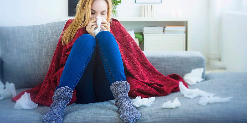 Woman at home on sofa wrapped in blanket blowing her nose