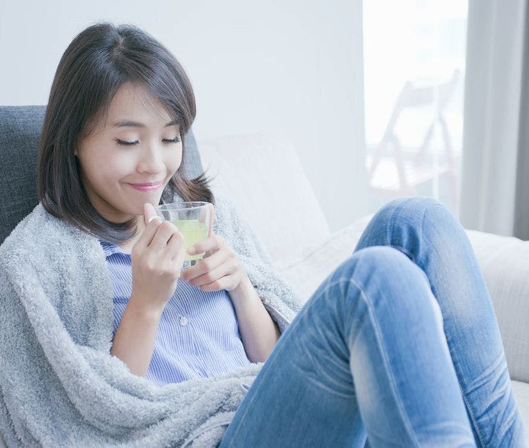 Une jeune femme noire est assise sur un canapé, recroquevillée sur elle-même et enveloppée d’une couverture. Elle présente possiblement des symptômes de rhume ou de la grippe.