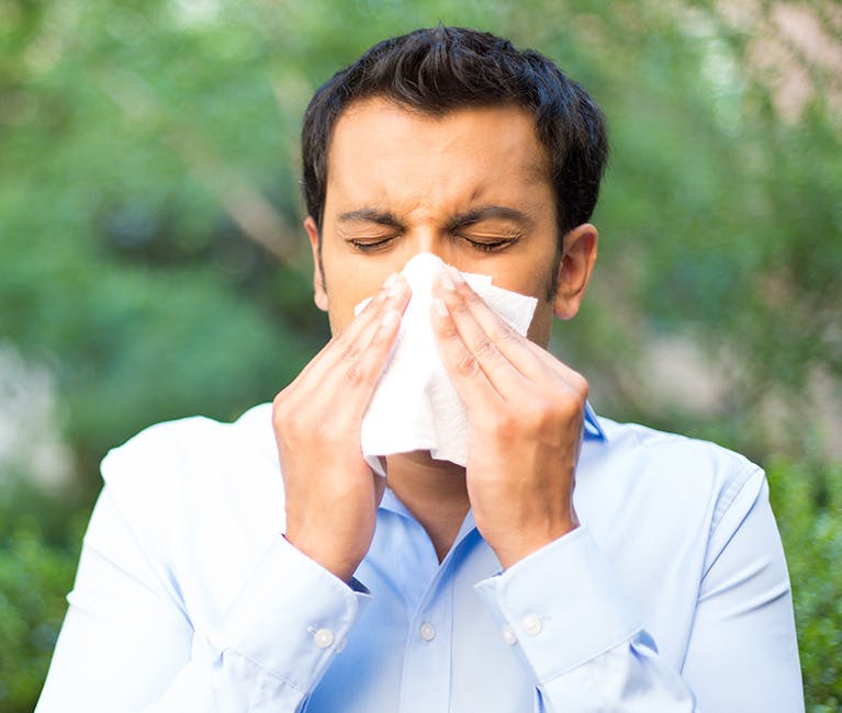 A black-haired man in a blue shirt is trying to blow his nose in a tissue