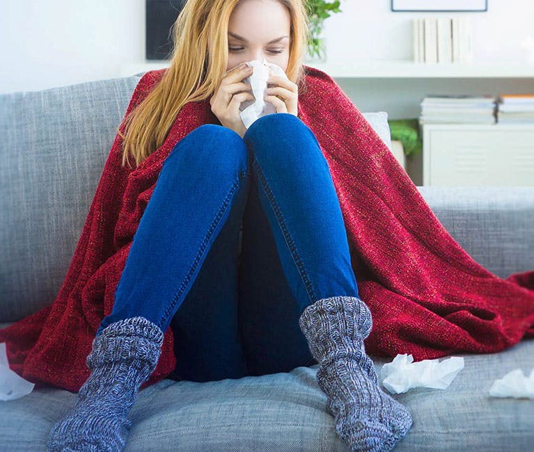 Woman at home on sofa wrapped in blanket blowing her nose