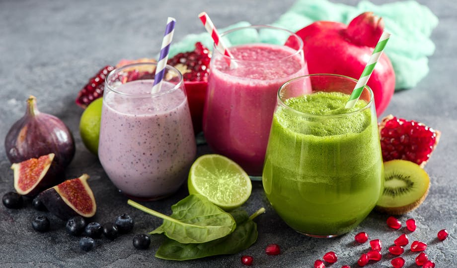 Three different smoothies – in purple, red, and green – side by side, with fruits and vegetables next to them 