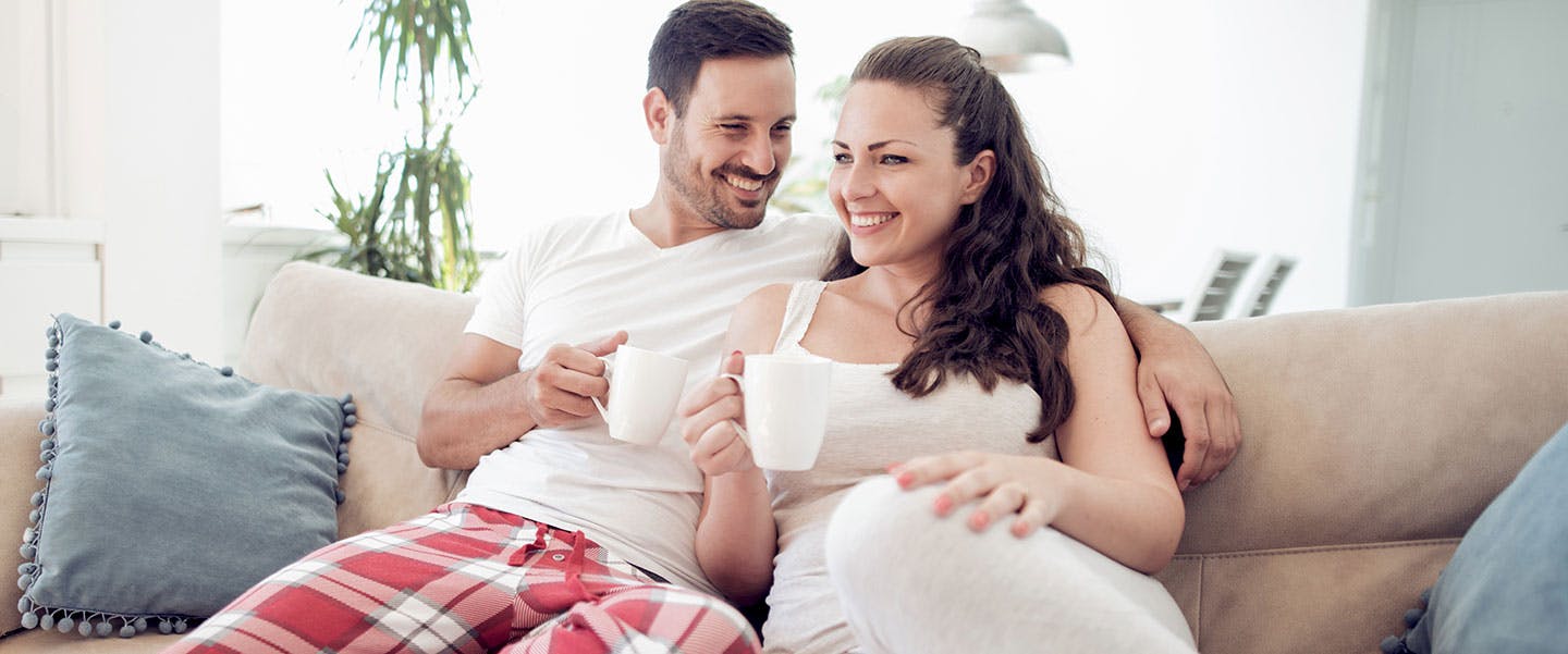 Happy couple in pajamas relaxes on couch with warm drinks in the morning