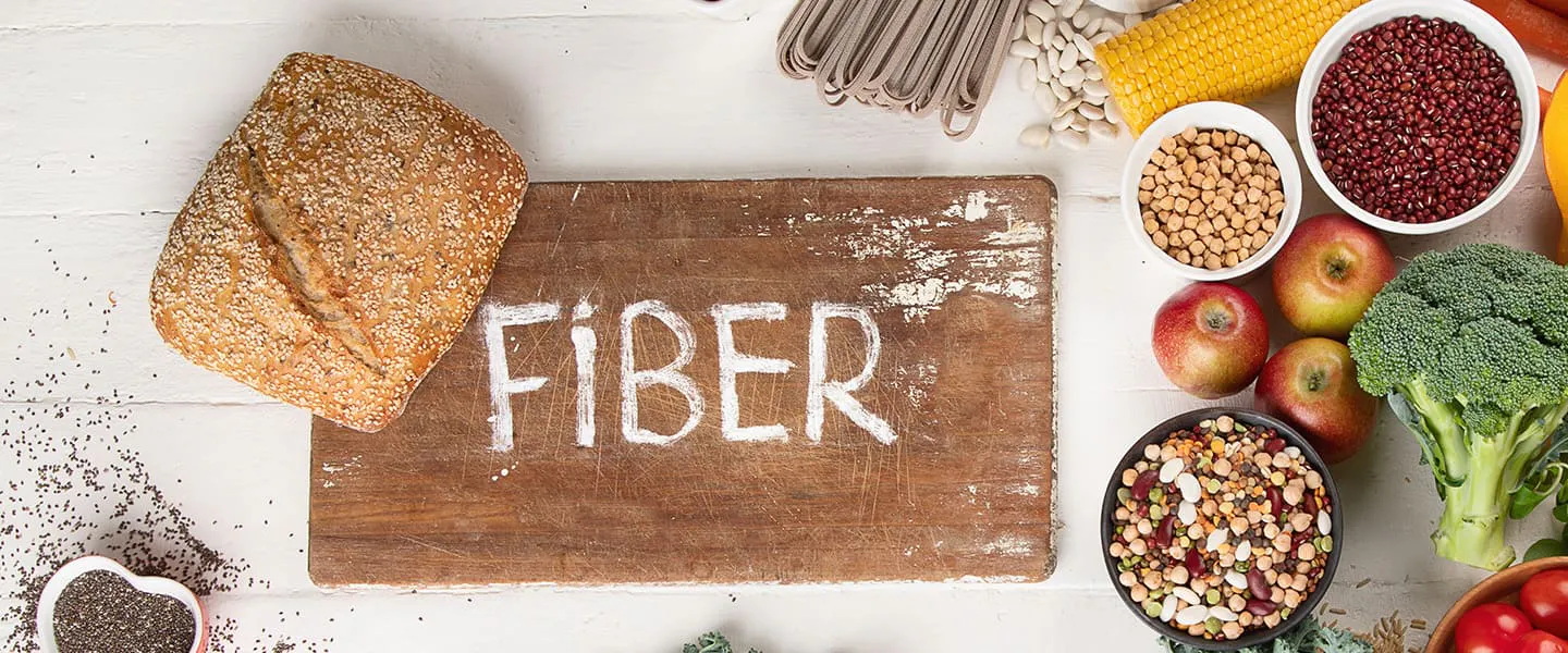 Foods high in fiber on a white background, including bread, chia seeds, nuts, beans, broccoli, apples, and greens.
