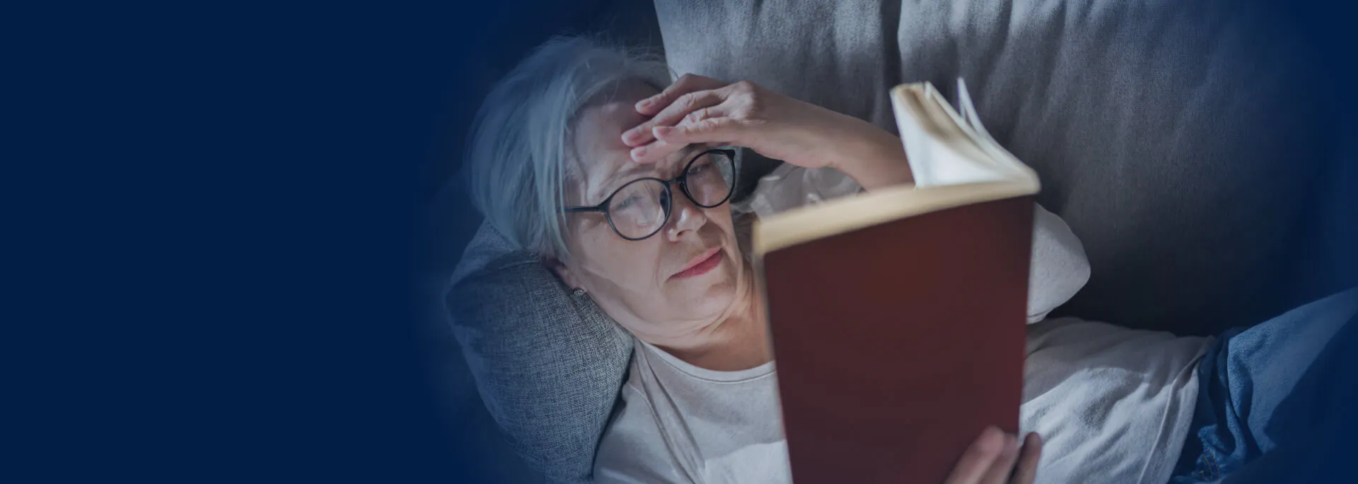 An older person lying on a couch, holding their head with one hand and reading a book.