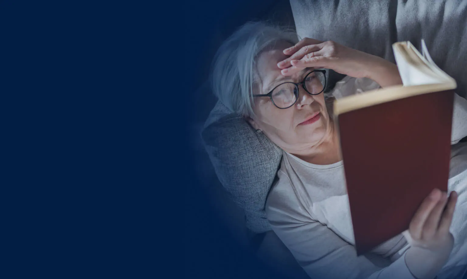 An older person lying on a couch, holding their head with one hand and reading a book.