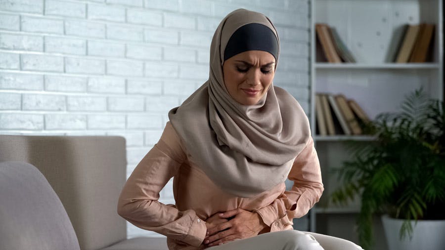Young woman wearing a headscarf hunched over on the couch dealing with period cramps
