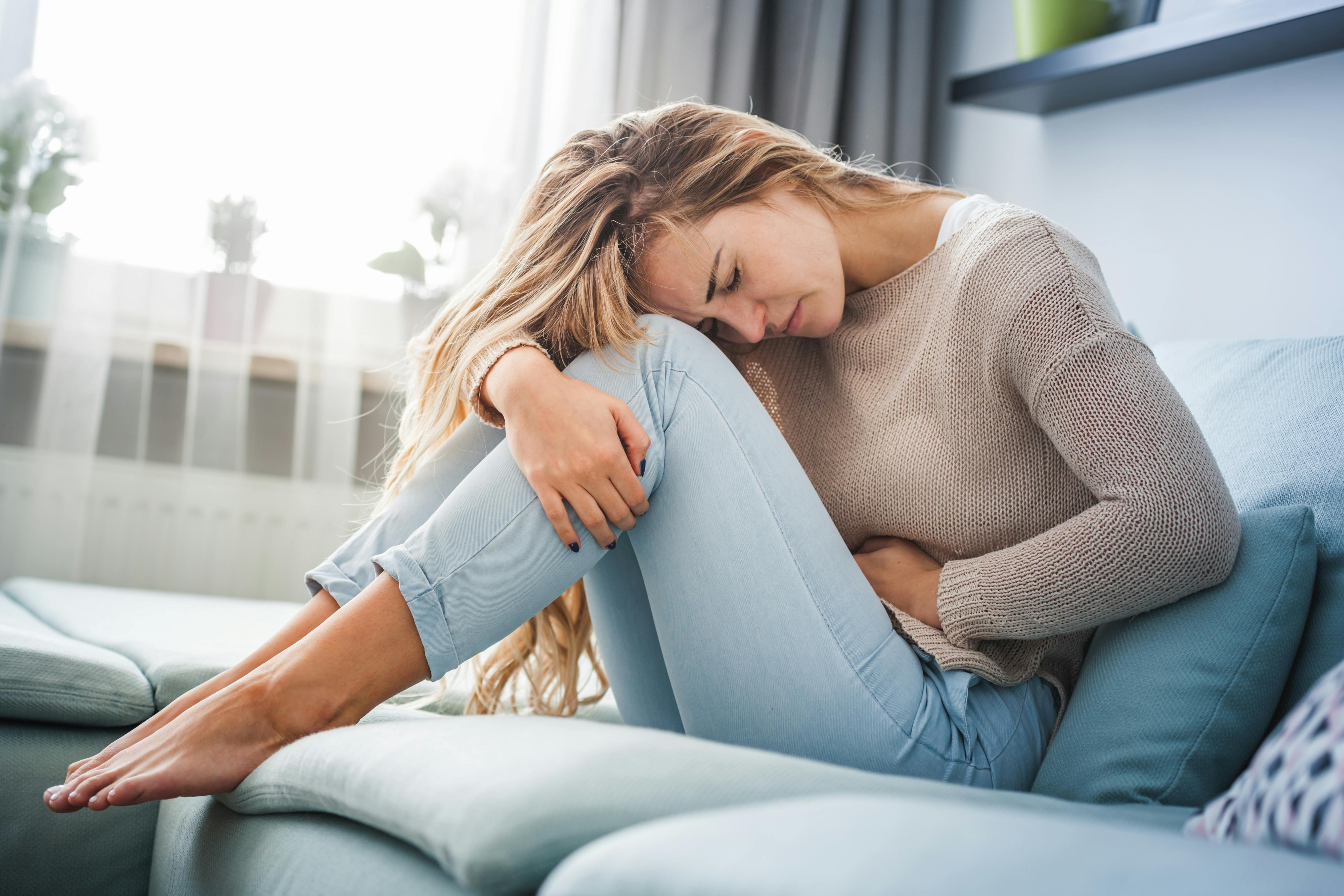 Young blonde woman hunched over with abdominal pain sitting on the sofa at home