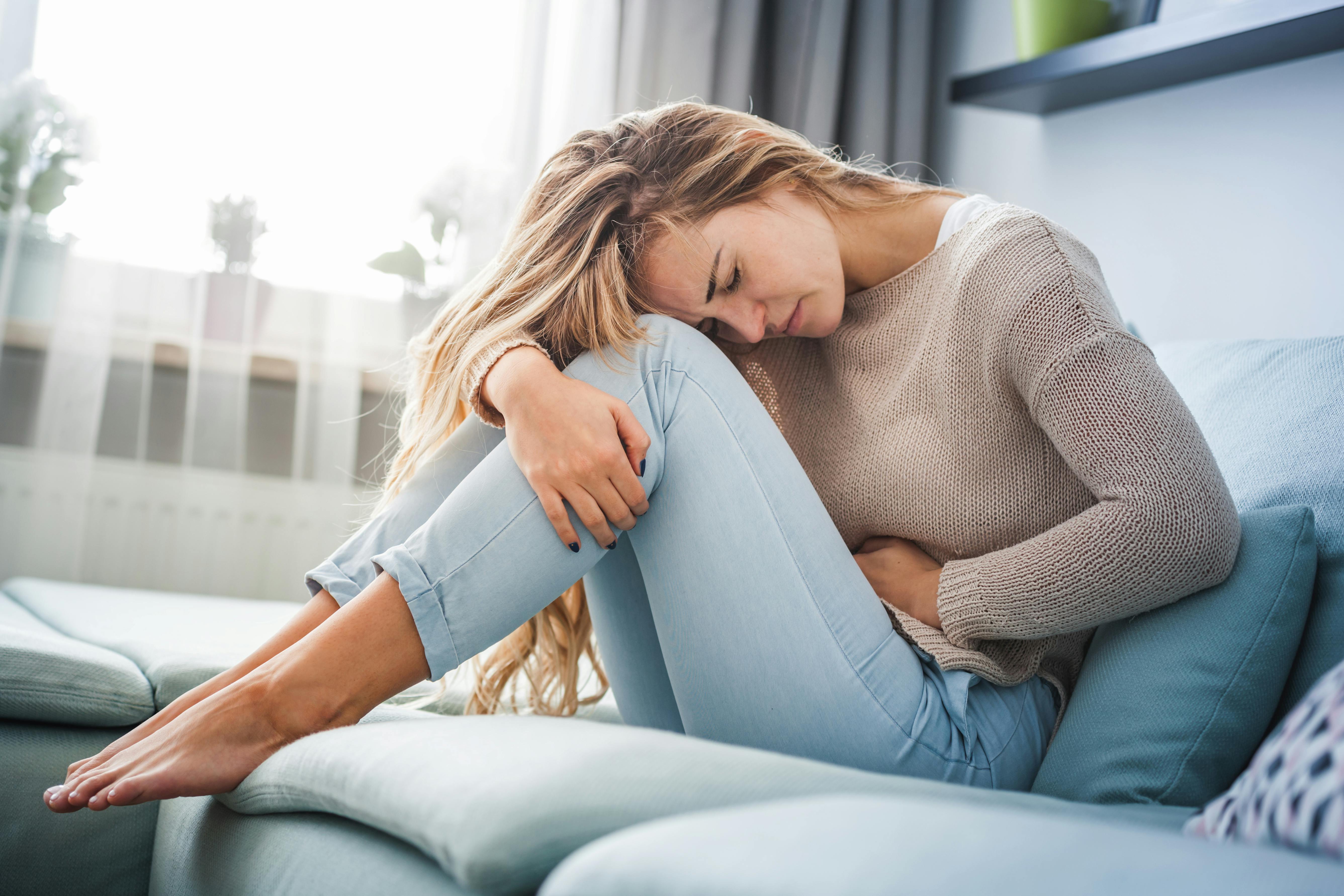 Young blonde woman hunched over with abdominal pain sitting on the sofa at home Young blonde woman hunched over with abdominal pain sitting on the sofa at home