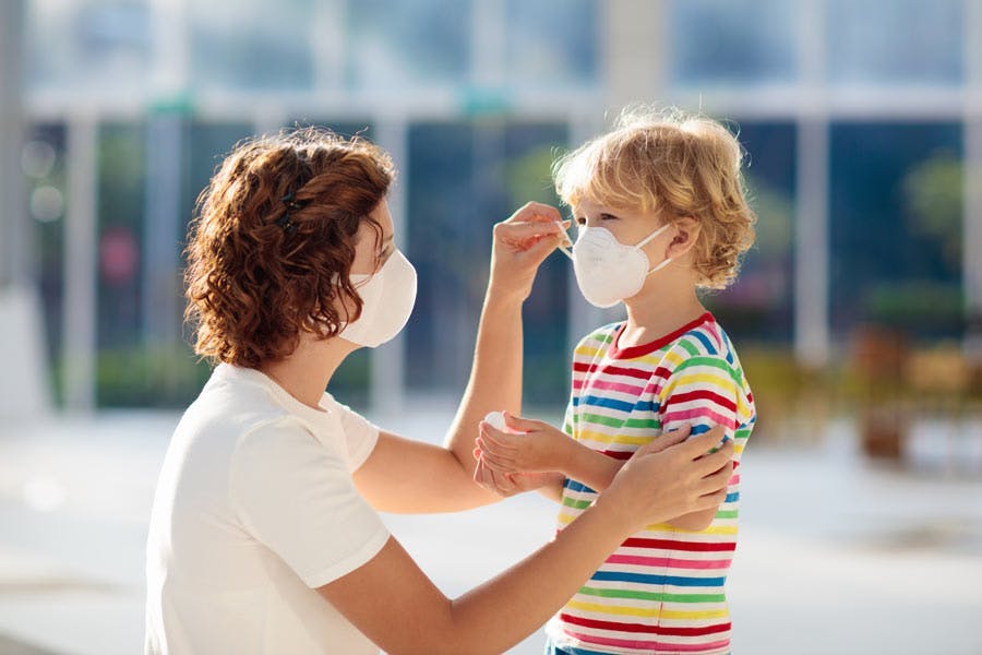 Mother wearing a face mask leaning down to help her young child put on a face mask