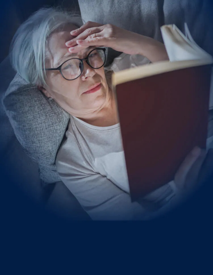 Personne âgée couchée sur un canapé, se tenant la tête d’une main et lisant un livre
