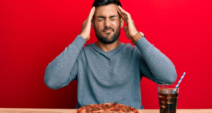 Hombre se toma la cabeza en señal de dolor al frente de él una pizza y una gaseosa