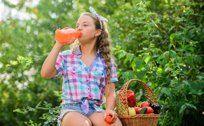 Girl drinking fruit juice next to a basket of vegetables