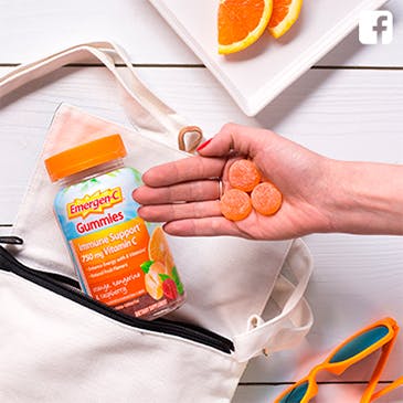 Hand holding three Emergen-C Gummies with bottle laying beside sunglasses and orange slices.