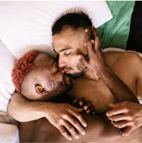 Two men in bed, one kissing the other's cheek tenderly, both shirtless and intimate