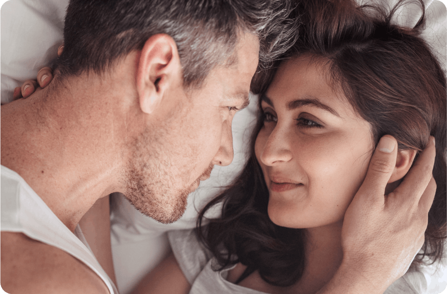 A couple lying in bed, smiling at each other, the man gently touching the woman’s face.