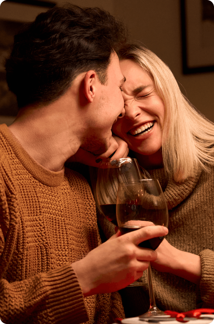 A couple laughing and enjoying a glass of wine together at a cozy, dimly lit setting.