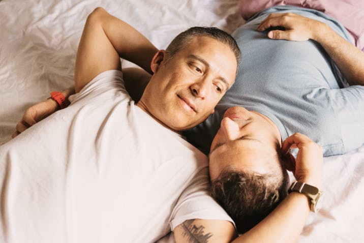 Two men lying on a bed, smiling and gazing at each other