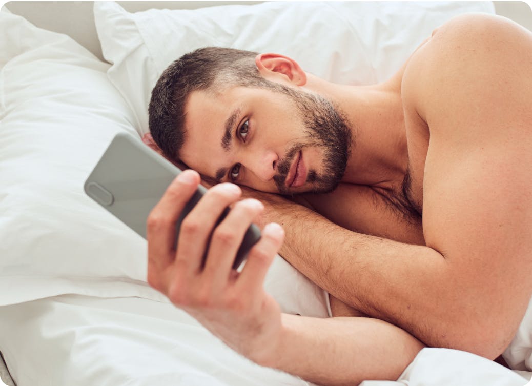 A man lying in bed, looking at his smartphone with a thoughtful expression.