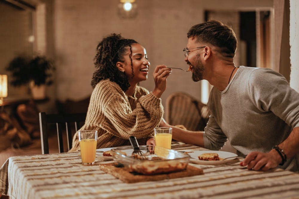 A joyful couple dining together; a woman feeds a man across the table in a warmly lit room, both smiling and engaged