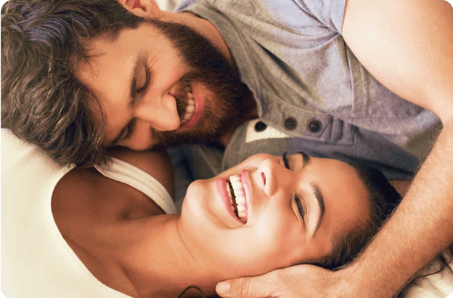 Bearded man in a gray shirt, smiling widely as he lies on top of a laughing woman in bed