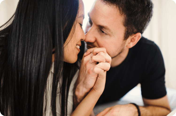Couple close together in bed, woman smiling as man kisses her hand gently