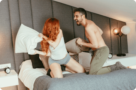 A couple having a playful pillow fight on a bed, both laughing and enjoying the moment.