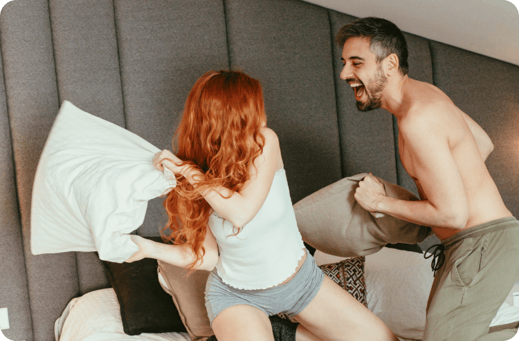 A joyful pillow fight between a man and a woman on a bed, both laughing in a playful and lighthearted moment
