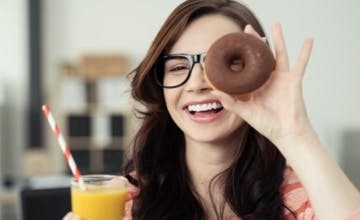 young brunette woman with black plastic glasses holding a chocolate donut and a glass of orange juice