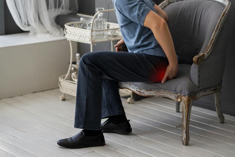 man in blue jeans sitting on gray chair rubbing buttocks in pain