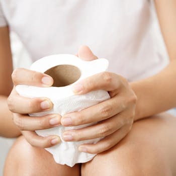 Person experiencing constipation sits on a toilet, holding a roll of toilet paper