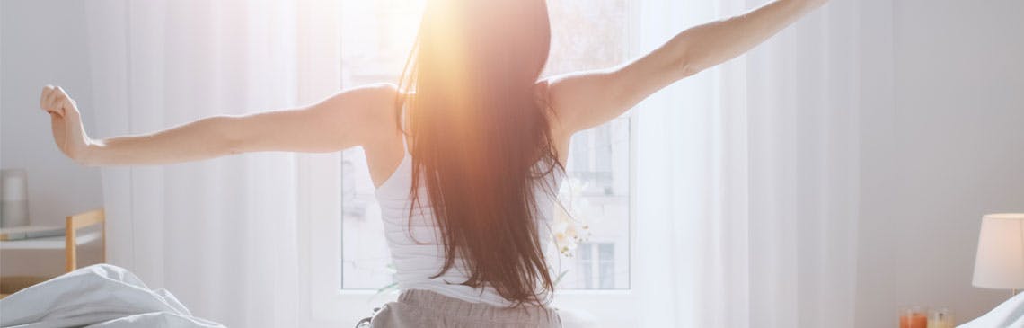 Woman stretches in bed and looks out a sunny window in the morning