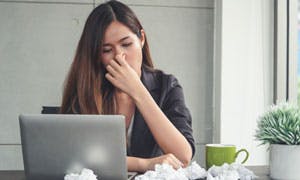 A woman sitting at a table and squeezing her nose with used balls of tissues, a mug and a laptop on the table