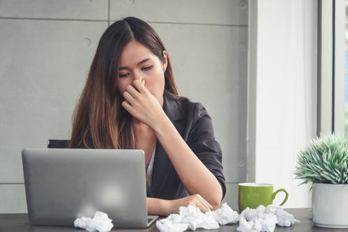 A woman sitting at a table and squeezing her nose with used balls of tissues, a mug and a laptop on the table