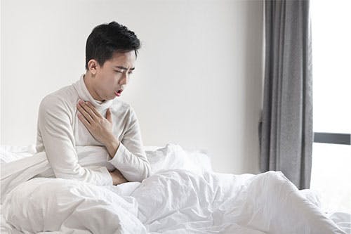 Man sitting on a bed while coughing and holding his chest Man sitting on a bed while coughing and holding his chest