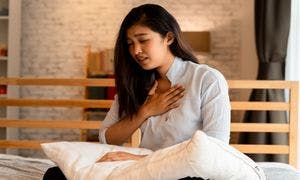 Portrait of a young adult Asian woman sitting in bed in a bedroom with a hand on her chest to signify having difficulty breathing due to chest congestion.