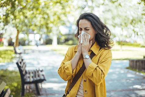 Brunette woman in a yellow jacket is blowing her nose, unsure whether she has an allergy or cold.
