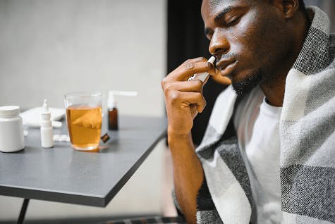 Man wearing a blanket and surrounded by various cold and flu medicines and using a nasal spray to help his nasal congestion