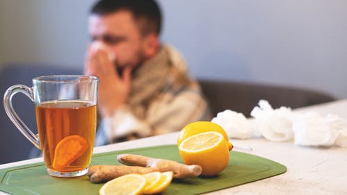 A cup of ginger tea and lemons with a person blowing their nose in the background