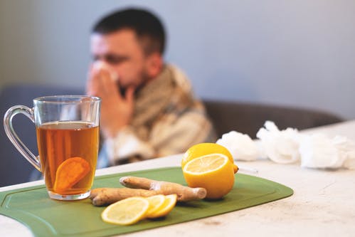 A cup of ginger tea and lemons with a person blowing their nose in the background