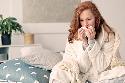 A woman is sitting up on bed, wrapped in a blanket and holding a tissue to her face