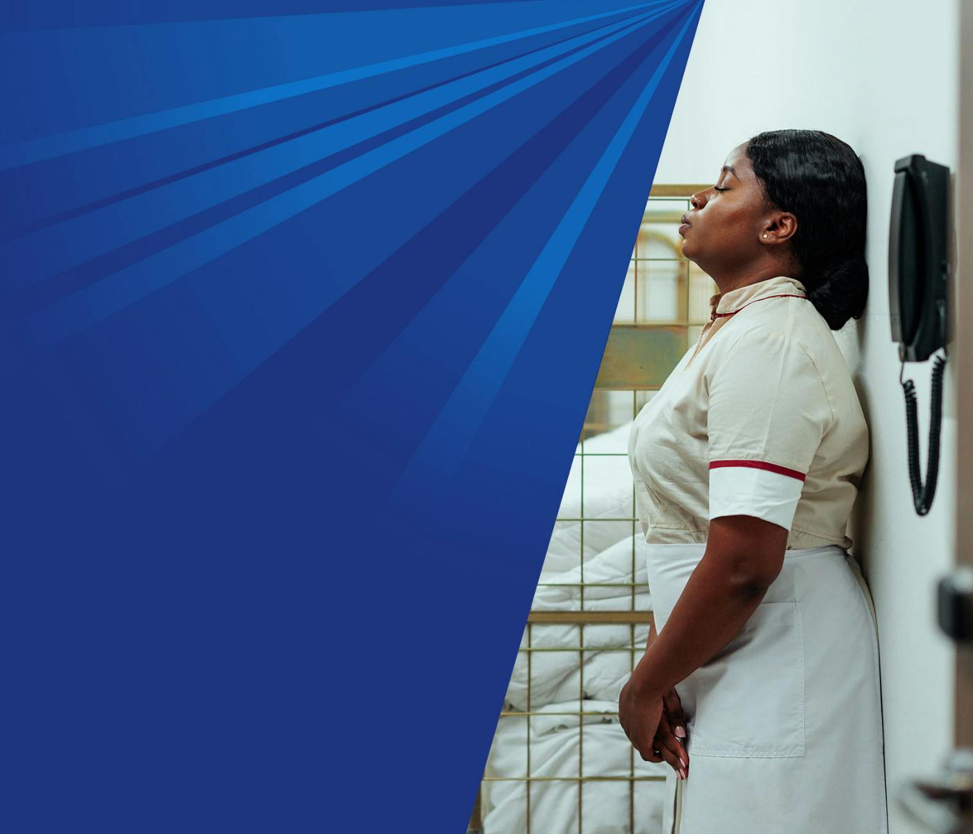 A woman in hotel cleaning staff attire leans against the wall in a large laundry room, resting her eyes. 