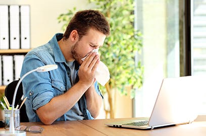 A man sitting at a desk is blowing his nose