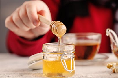 Hand dipping into honey with wooden stick, tea in background