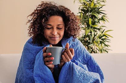 A woman wrapped in a blanket is holding a mug of hot liquid. There is steam rising from the cup.