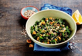 A bowl of kale salad with mixed grains and nuts sits on a table with a blue napkin, there is a lemon off to the right hand side