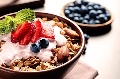 A bowl of yoghurt with strawberries, blueberries and nuts with a sprig of mint sits on a surface, there is a bowl of blueberries in the background