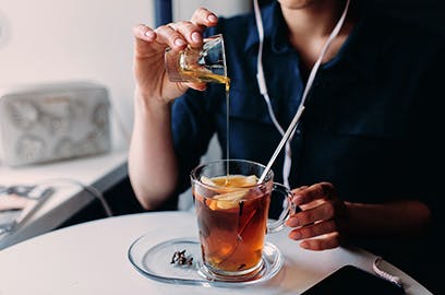 A person sitting at a table is pouring honey into hot tea with a lemon slice in it