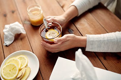 Hands are resting on a table holding a glass mug with a lemon slice in it. Other lemon slices sit on a plate off to the side with a tissue box.